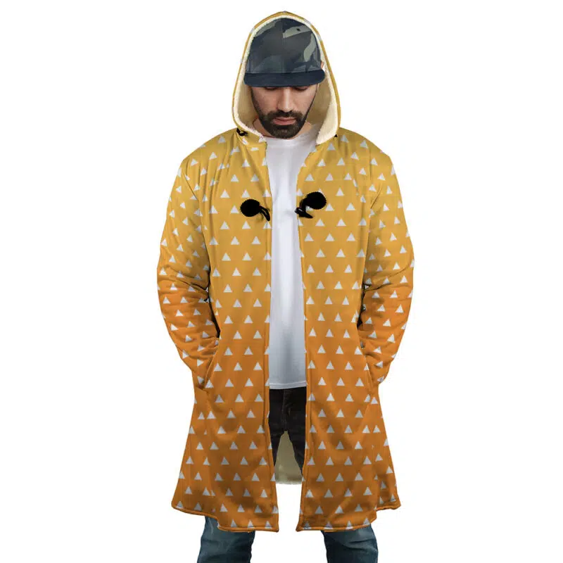 A man stands with hands in pockets, looking down, wearing the Zenitsu Agatsuma Demon Slayer Dream Cloak Coat—a hooded yellow and orange coat with white triangles and black crow silhouettes—over a camouflage cap and white T-shirt.