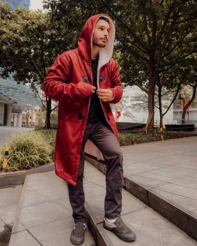 A man wearing the Edward Elric Fullmetal Alchemist Dream Cloak Coat stands on a city sidewalk, looking to the side. He pairs the long red hooded coat with black buttons with dark pants and black shoes, surrounded by trees and buildings.