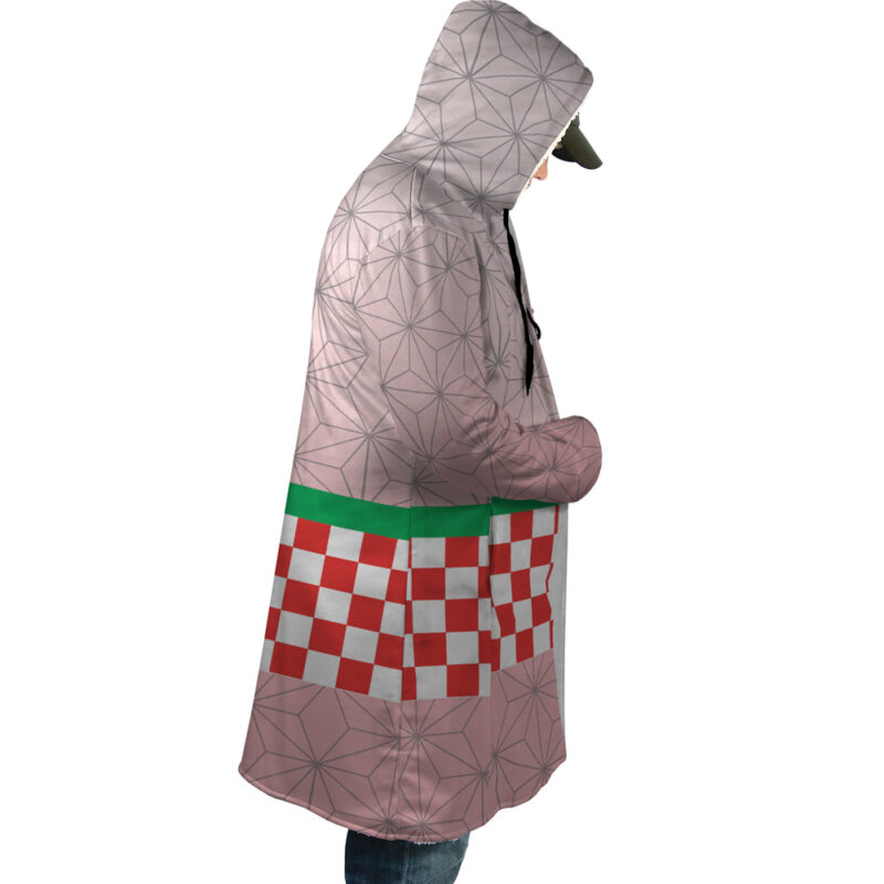 A person stands in profile, hands in pockets, wearing jeans, a cap, and the Nezuko Kamado Demon Slayer Dream Cloak Coat featuring geometric patterns, a green stripe, and a red-and-white checkerboard design.