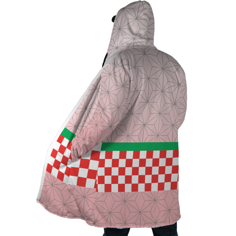 Someone wears the Nezuko Kamado Demon Slayer Dream Cloak Coat, which features a geometric pattern and a red-and-white checkerboard stripe with a green border near the bottom, set against a pale pink background with subtle lines.