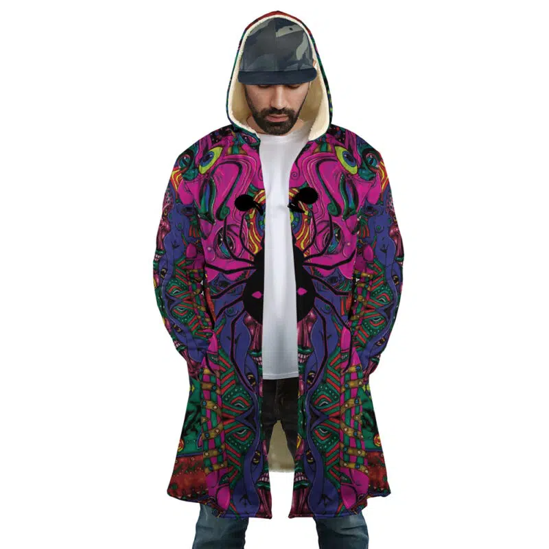 A man wearing the Trippy Mystic Faces Hisoka Hunter X Hunter Dream Cloak Coat, featuring colorful psychedelic patterns in pink, purple, green, and blue, stands with his hands in his pockets, paired with jeans and a camo cap.