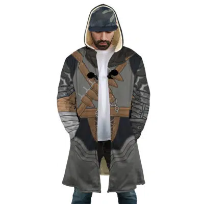 A man wears the Guts Armor Berserk Dream Cloak Coat, designed with medieval armor-inspired prints, including straps, buckles, and metallic patterns. He pairs it with a baseball cap and looks down.