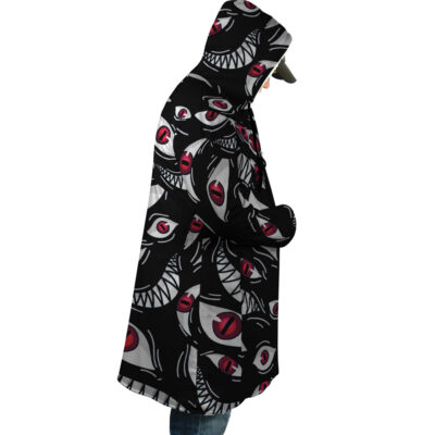 A person wears the Pride Fullmetal Alchemist Dream Cloak Coat, which features a bold black, red, and white eye-and-teeth pattern. Theyre facing sideways with hands in pockets, and a cap under the hood.