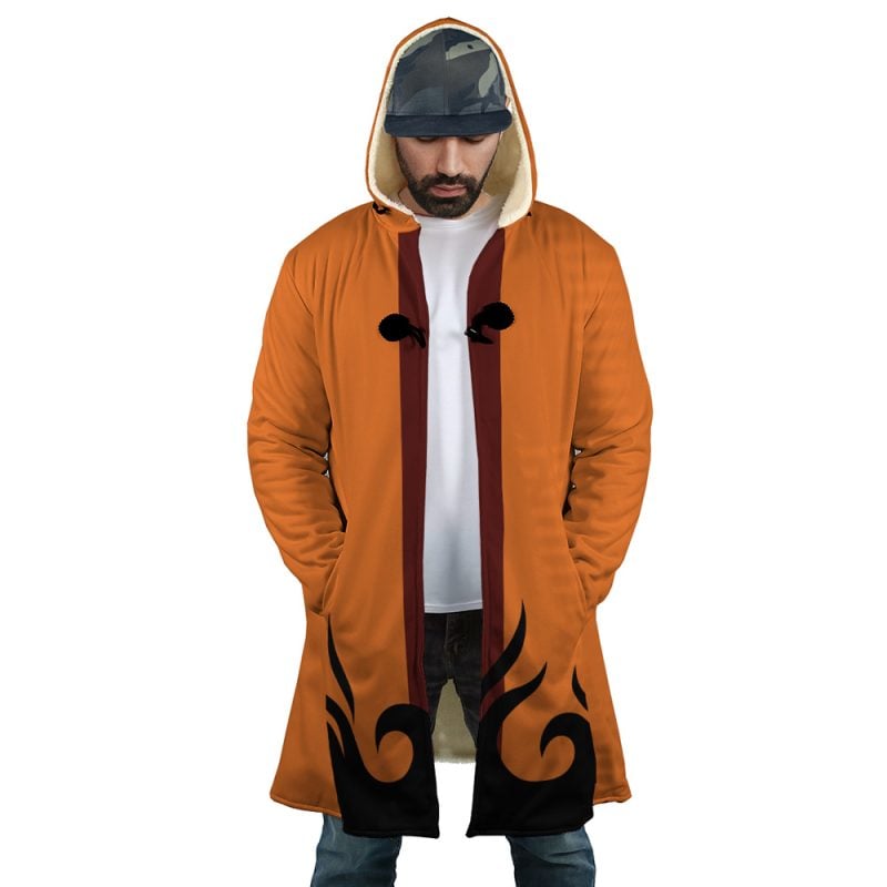 A man wears the Hokage Orange Shippuden Dream Cloak Coat, featuring black flame patterns near the hem and a cream lining, paired with a camo-print cap. He stands with hands in pockets, looking down.