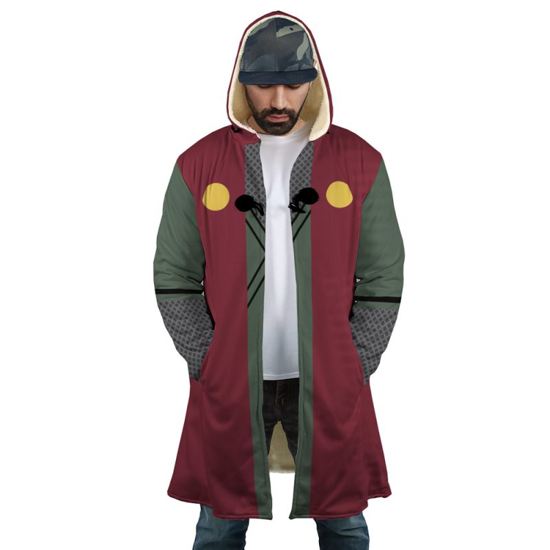 A man wearing the Jiraiya Uniform Shippuden Dream Cloak Coat, a long hooded coat with maroon, olive green, and gray panels, yellow accents, and black chest cords. He pairs it with a white t-shirt, blue jeans, and a patterned cap.