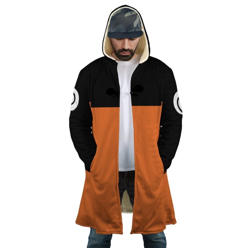 A man wears the Uzumaki Shippuden Dream Cloak Coat—long, hooded, orange and black with white circular sleeve designs—paired with a white t-shirt, blue jeans, and a camouflage cap. He looks down with hands in his pockets.