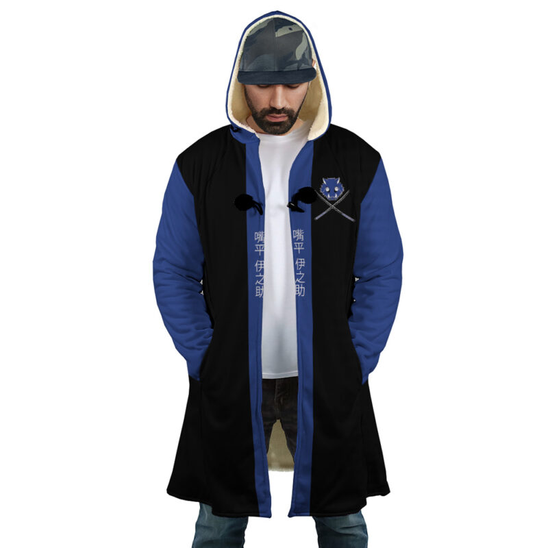 A man wearing the Hashibira Inosuke Demon Slayer Dream Cloak Coat, a blue and black hooded coat with fur lining, crossed swords and a crest on the chest, and white Asian script down the front. He looks down while wearing a cap.