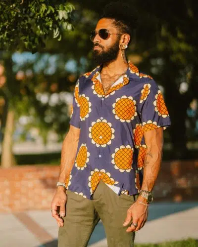 A bearded man in sunglasses stands outdoors wearing the Navy Blue Monkey D. Luffy Lucy Dressrosa One Piece Button Up Hawaiian Shirt. Trees and a brick wall are visible in the background.