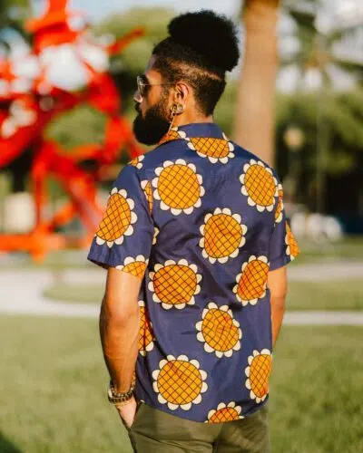 A man with a beard and dark hair in a high puff stands outdoors wearing the Navy Blue Monkey D. Luffy Lucy Dressrosa One Piece Button Up Hawaiian Shirt, sunglasses, and earrings. Blurred greenery and a red sculpture are visible behind him.