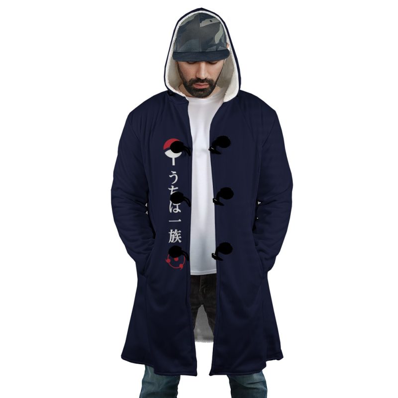 A man stands with hands in pockets, looking down, wearing the Uchiha Clan Sharingan Shippuden Dream Cloak Coat—a dark blue hooded coat with Japanese characters and red-and-white fan symbols—over a white shirt and printed cap.