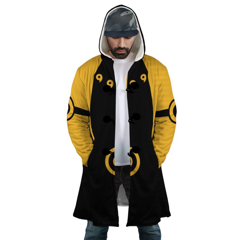 A man wears the Six Paths Sage Mode Shippuden Dream Cloak Coat, a long hooded coat with yellow and black circular and comma-like designs. He stands with hands in pockets, looking down, wearing a gray cap and white shirt underneath.