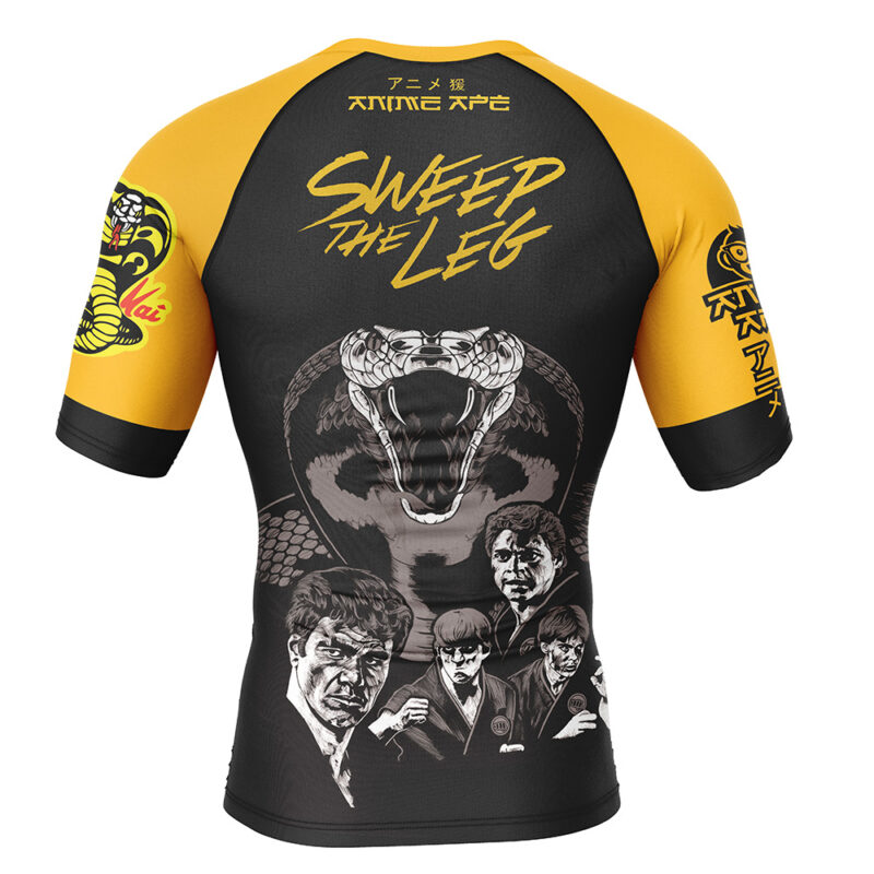Back view of the Sweep the Leg Cobra Kai Short Sleeve Rash Guard Compression Shirt in black and yellow, with a large cobra, four illustrated men, Japanese text, "SWEEP THE LEG", and sleeve designs including the Cobra logo.