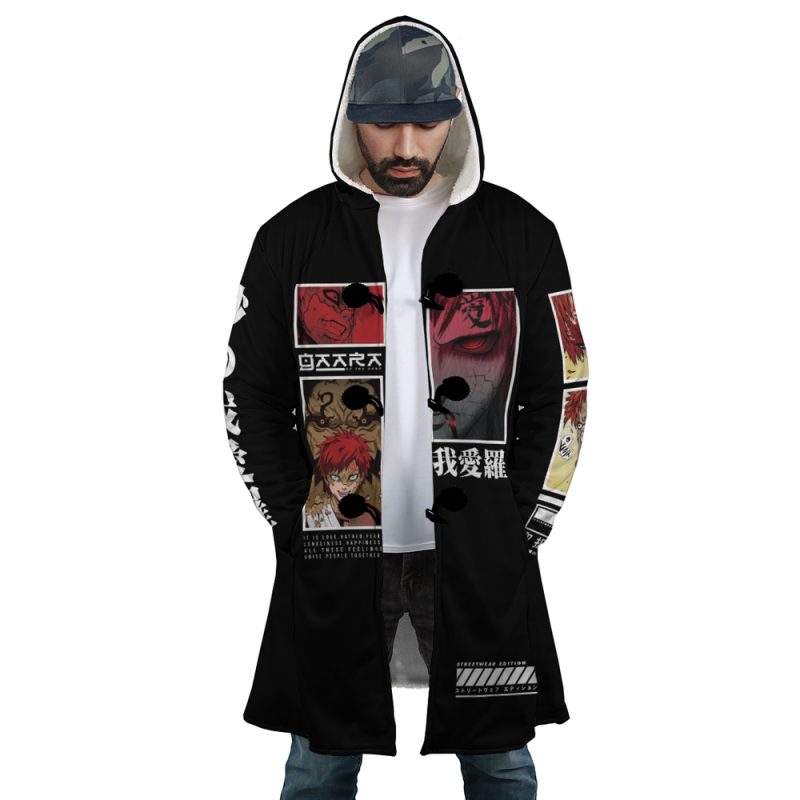 A man wears the Gaara Shippuden Dream Cloak Coat, a long black hooded coat with anime-style graphics and Japanese text in red, white, and black. The design features illustrated faces and dramatic scenes as he looks down with hands in his pockets.