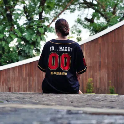 A person with tied-back hair sits on stone steps, wearing the Central City Chimeras Fullmetal Alchemist Baseball Jersey (black, ED...WARD? 00, Japanese text). Lush trees and a curved wooden wall are in the background.