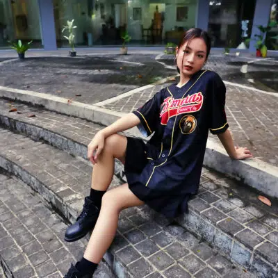A young woman sits on outdoor stone steps wearing the Central City Chimeras Fullmetal Alchemist Baseball Jersey and black boots. She looks at the camera with her hair tied back, surrounded by green plants and a modern building in the background.