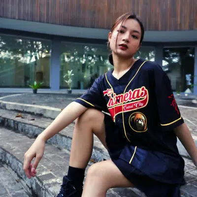 A young woman sits confidently on outdoor stone steps, wearing a black Central City Chimeras Fullmetal Alchemist Baseball Jersey, with one knee up and her arm resting casually on her leg as she looks at the camera.