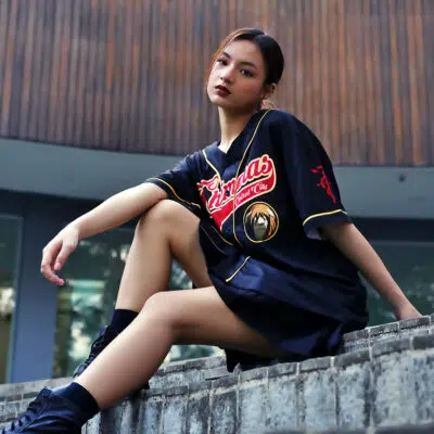 A young woman sits on a concrete ledge outside, confidently wearing the Central City Chimeras Fullmetal Alchemist Baseball Jersey with bold black, red, and gold accents and black boots. A curved wooden wall is in the background.