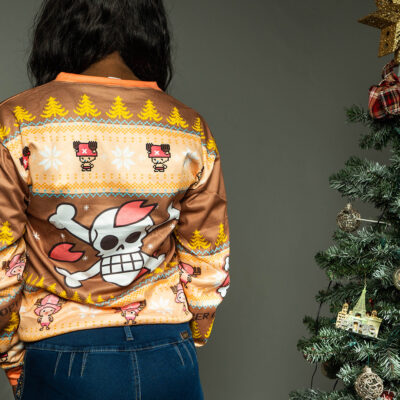 A person stands with their back to the camera, wearing the Christmas Tony Chopper One Piece Ugly Christmas Sweater, which features cartoon skulls, crossbones, holiday trees, and snowflakes. A decorated Christmas tree appears on the right.