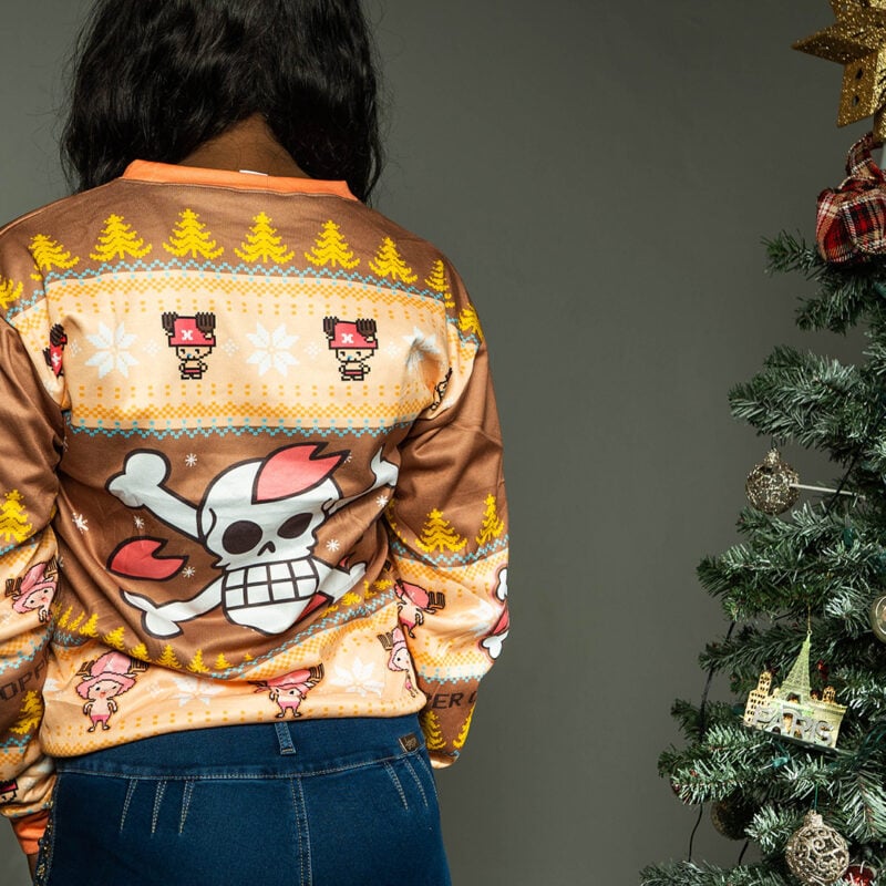 A person stands with their back to the camera, wearing the Christmas Tony Chopper One Piece Ugly Christmas Sweater, which features cartoon skulls, crossbones, holiday trees, and snowflakes. A decorated Christmas tree appears on the right.
