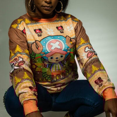 A woman wearing the Christmas Tony Chopper One Piece Ugly Christmas Sweater, adorned with festive and cartoon Chopper designs, squats with her hands on her knees against a neutral background.