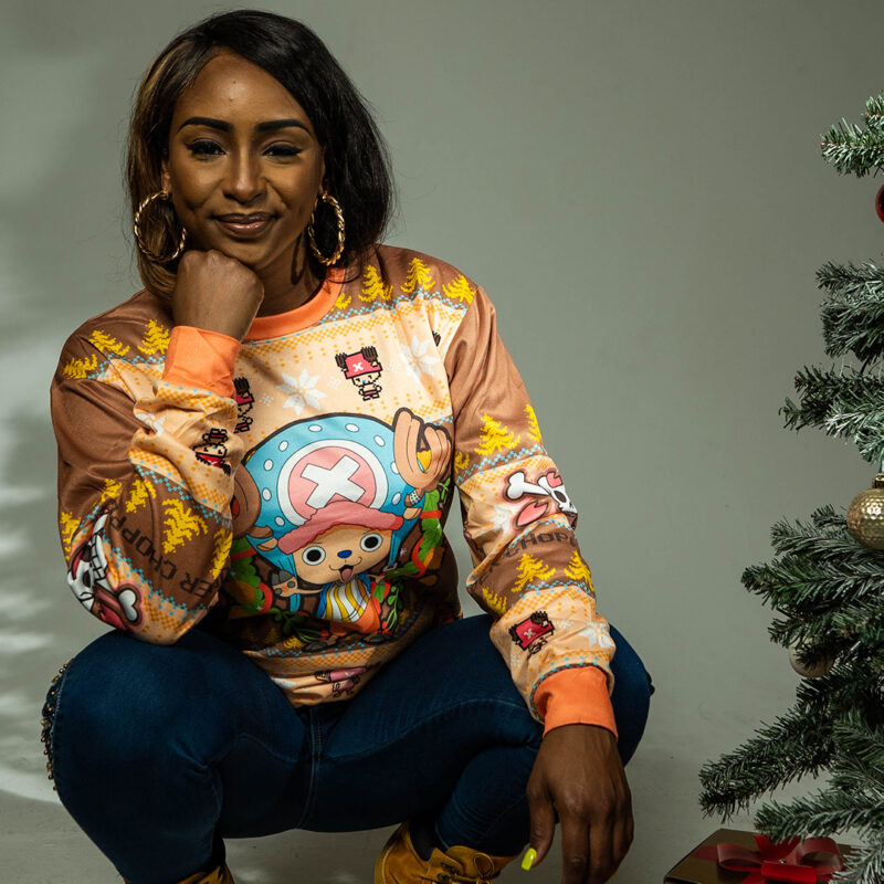 A woman with long dark hair, hoop earrings, and a Christmas Tony Chopper One Piece Ugly Christmas Sweater crouches and smiles next to a decorated Christmas tree with ornaments.