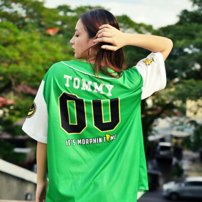 A person with long hair stands outdoors, wearing the Green Dragonzords Tommy Oliver Power Rangers Baseball Jersey featuring TOMMY 00 and ITS MORPHIN TIME on the back, surrounded by trees and blurred buildings.