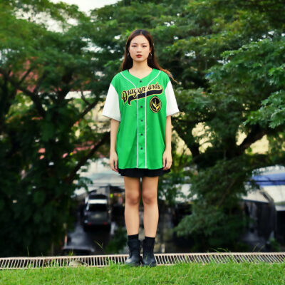 A young woman stands outdoors wearing a Green Dragonzords Tommy Oliver Power Rangers Baseball Jersey, paired with a black skirt and boots, with trees and parked cars visible in the background.