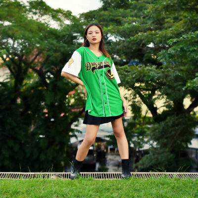 A young woman stands confidently outdoors in a Green Dragonzords Tommy Oliver Power Rangers Baseball Jersey, paired with a black skirt and ankle boots, with lush green trees in the background.