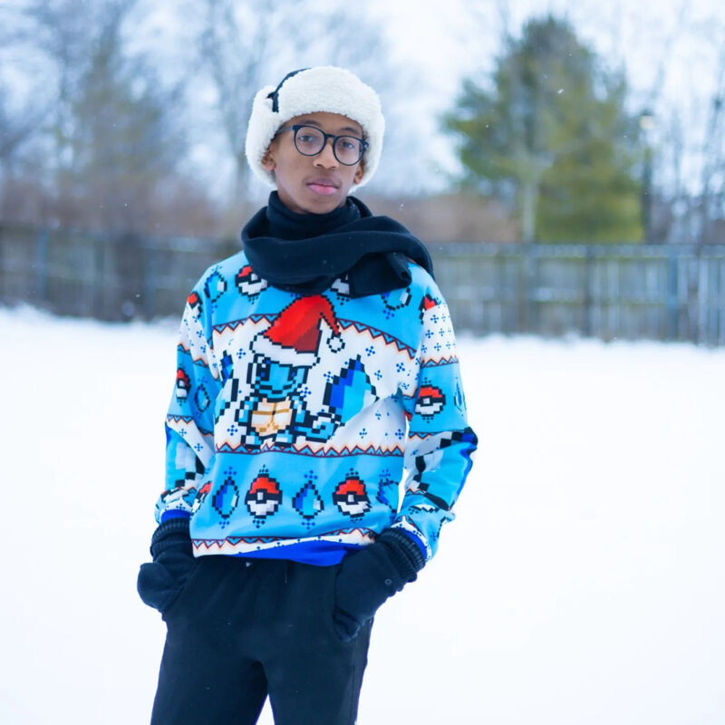 A person stands outside in the snow wearing a white winter hat, glasses, black scarf and gloves, and a blue Pokemon Squirtle Ugly Christmas Sweater with a festive design. Trees and a fence are visible in the background.