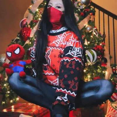 Wearing a Spiderman Uniform Ugly Christmas Sweater and red mask, a person sits cross-legged in front of a decorated Christmas tree, holding a Spider-Man plush toy. Wrapped presents are under the tree in the background.