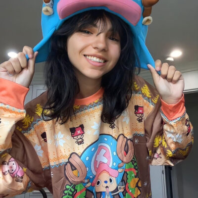 A young woman with dark hair smiles while wearing the Christmas Tony Chopper One Piece Ugly Christmas Sweater, featuring a colorful, cartoon Tony Chopper design. She stands indoors under bright lighting and holds the sides of a large blue character hat.
