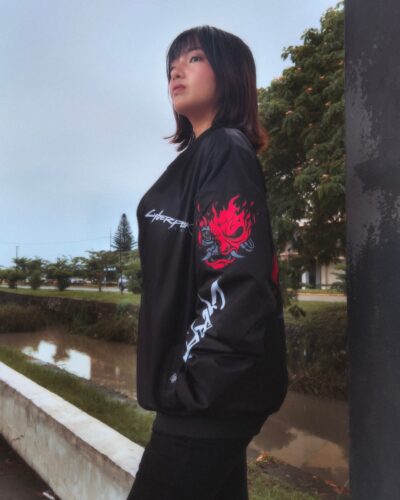 A woman with shoulder-length dark hair stands outdoors, wearing the Johnny Silverhand Cyberpunk 2077 Casual Bomber Jacket, which features red and white graphics like a flaming skull on the sleeve. She gazes upward with trees and water behind her.