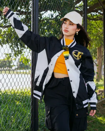 A young woman poses outdoors in a Black Ranger Mighty Morphin Power Rangers Varsity Jacket, yellow top, and light cap near a wire fence with greenery and trees in the background on a sunny day.