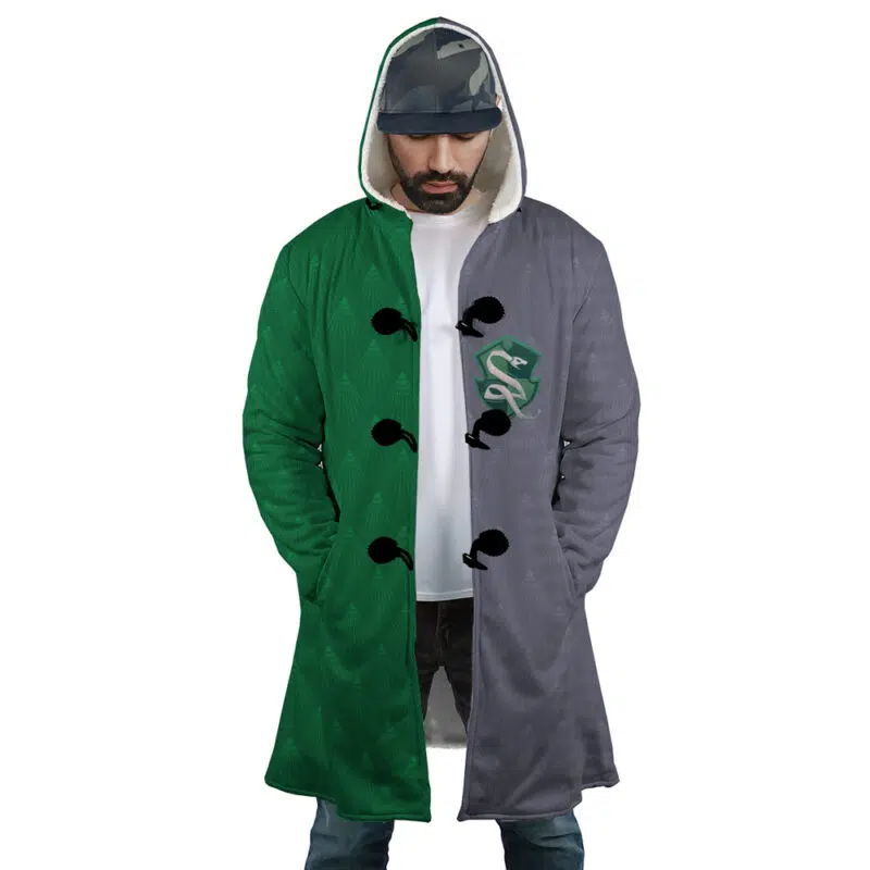 A man wears the Hogwarts Legacy Slytherin Harry Potter Dream Cloak Coat—long, hooded, split green and gray with a snake crest—standing hands in pockets, looking down.