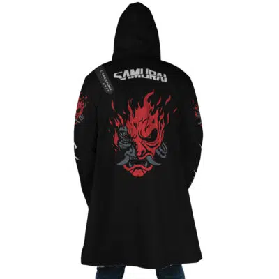 A person wearing the Johnny Silverhand Cyberpunk 2077 Dream Cloak Coat, a long black hooded coat with SAMURAI and a large red fiery demon face graphic on the back, stands facing away from the camera.