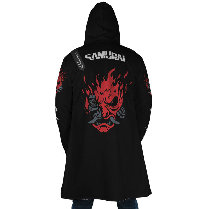A person wearing the Johnny Silverhand Cyberpunk 2077 Dream Cloak Coat, a long black hooded coat with SAMURAI and a large red fiery demon face graphic on the back, stands facing away from the camera.