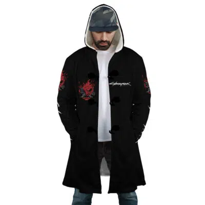A man wearing the Johnny Silverhand Cyberpunk 2077 Dream Cloak Coat with red skull designs and Cyberpunk text, a white T-shirt, blue jeans, and a camo cap stands facing forward with his hands in his coat pockets.