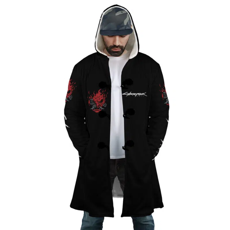 A man wearing the Johnny Silverhand Cyberpunk 2077 Dream Cloak Coat with red skull designs and Cyberpunk text, a white T-shirt, blue jeans, and a camo cap stands facing forward with his hands in his coat pockets.
