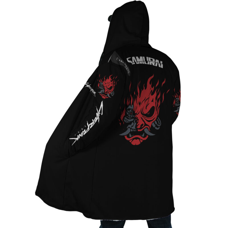 A person wearing the Johnny Silverhand Cyberpunk 2077 Dream Cloak Coat, a black hooded jacket featuring a large red and black flaming samurai mask and SAMURAI on the back, with extra artwork and text on the sleeves.