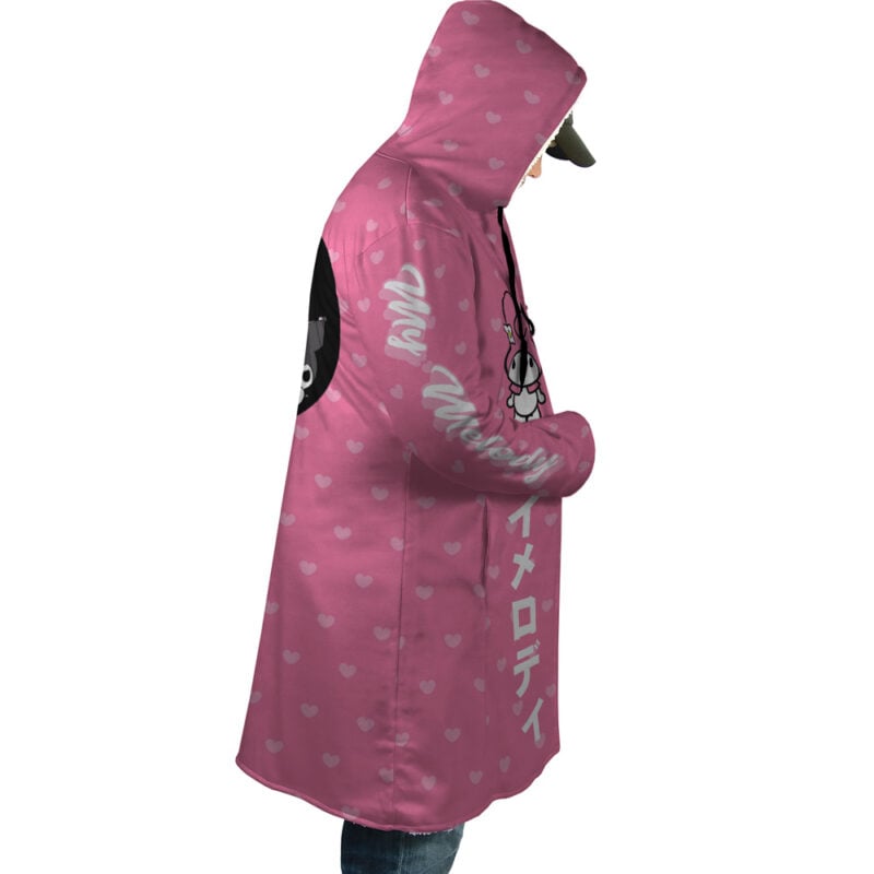 A person is wearing the My Melody and Kuromi Sanrio Dream Cloak Coat, featuring pink fabric with small white hearts, My Melody graphics, and white Japanese text on the sleeve and front, facing sideways with hands in pockets.