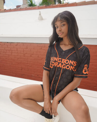 A young woman with long dark hair sits on a white rooftop in front of a red brick wall, wearing black shorts, a necklace, and a black Dungeons & Dragons Baseball Jersey with orange lettering and trim.