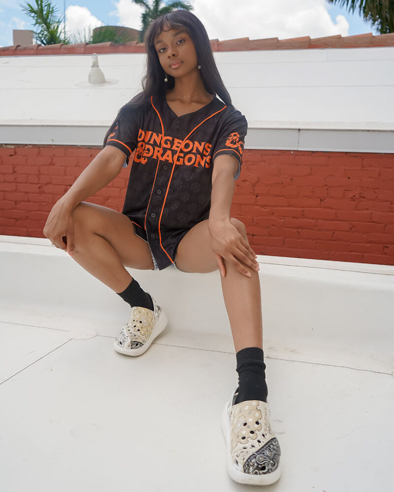 A person with long hair stands outdoors on a white surface, wearing a black Dungeons & Dragons Baseball Jersey, black shorts, patterned slip-on shoes, and black socks, looking confidently at the camera.