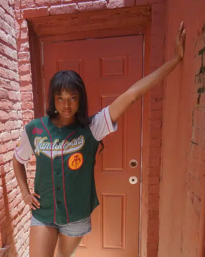 A woman poses confidently in a narrow brick alley, wearing the Mandalorians Boba Fett Star Wars Baseball Jersey and denim shorts, with one arm on the wall and a reddish-brown door behind her.