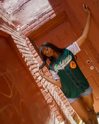 A woman stands in a narrow brick alleyway, wearing the Mandalorians Boba Fett Star Wars Baseball Jersey and denim shorts. She poses near a closed brown door, touching her hair as sunlight casts shadows on the scene.