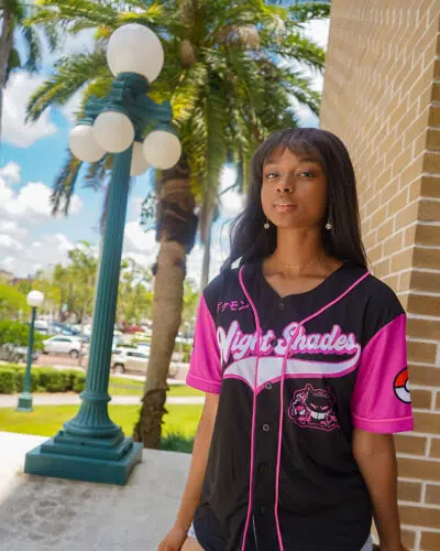 A woman stands outdoors by a brick wall and lamp post, wearing the Night Shades Ghost Type Pokemon Baseball Jersey in black and pink with a cat graphic. Palm trees, cars, and a bright blue sky appear in the background.