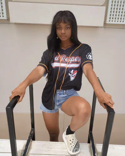 A woman wearing a Soul Reaper Ichigo Bleach Baseball Jersey, denim shorts, and white sneakers poses outdoors on stairs, holding the railing and looking at the camera.