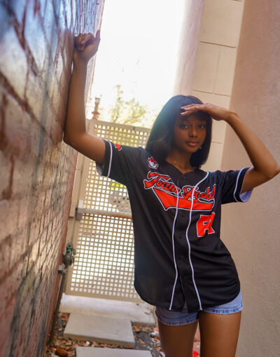 A young woman wearing a Team Rocket Grunt Black Pokemon Baseball Jersey and denim shorts stands in a narrow alley, leaning against a brick wall and shading her eyes from the sun with her hand.