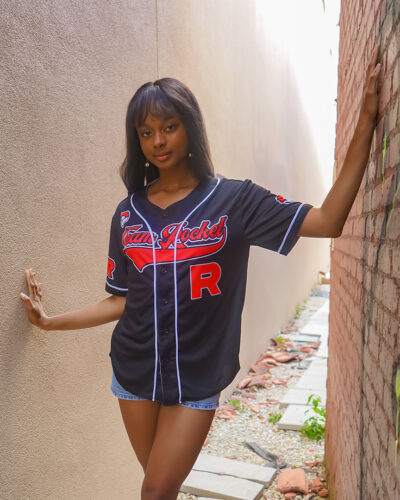 A woman wearing a Team Rocket Grunt Black Pokemon Baseball Jersey and denim shorts stands in a narrow outdoor walkway, resting one hand on each wall.