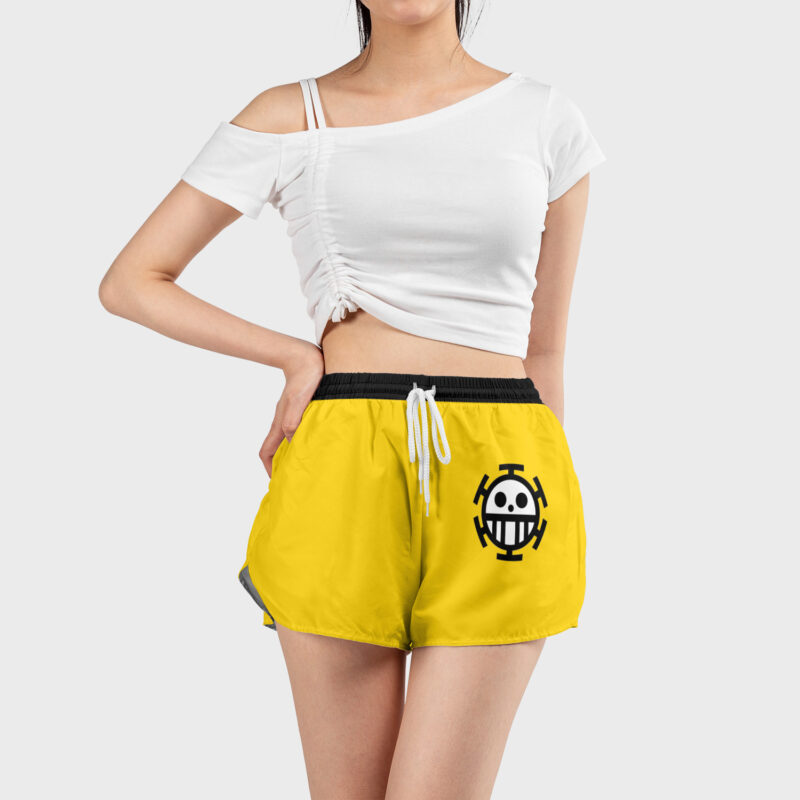 A woman stands against a plain light background wearing white off-shoulder crop top and Trafalgar Law One Piece Women's Board Shorts in yellow, featuring a black drawstring and a stylized smiley face logo on the left leg.