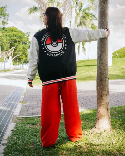 Someone wearing a Poke League Champion Pokemon Varsity Jacket and bright red pants stands on an outdoor path by grass and trees, facing away with one arm resting on a tree trunk.